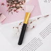 A gold and black silicone facial mask applicator brush resting on a pink paper background with dried floral elements.