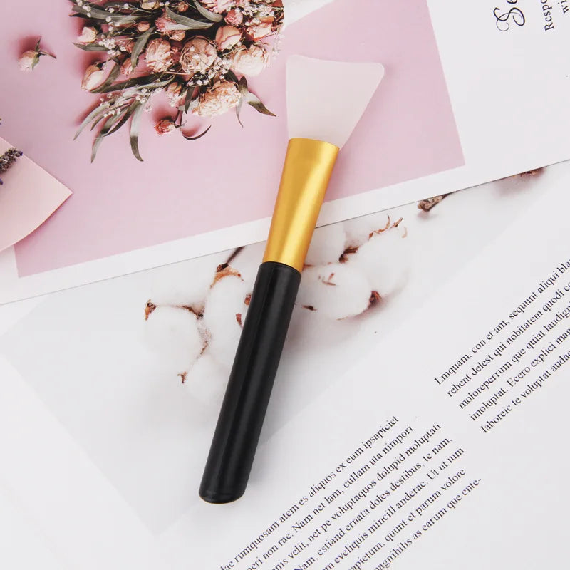 A gold and black silicone facial mask applicator brush resting on a pink paper background with dried floral elements.