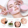 A woman applies a pink facial mask while a hand holds a silicone brush and other skincare products like liquid foundation and