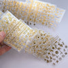 A hand holds a sheet of gold star nail art stickers for acrylic nails featuring various 3D decals and designs.