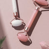 A set of pink and white resin facial massage rollers with rose gold handles resting on a light pink surface.