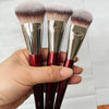 Trois pinceaux de maquillage à fondation liquides anglés avec ferrules argentées et poils roses tenus dans une main.
