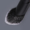 A close-up view of a black triangular precision concealer brush with white synthetic bristles on a gray surface.