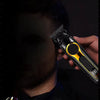 A close-up of a black electric hair clipper with a yellow accent, shown being held by a hand against a dark background.