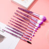 A set of ten professional makeup brushes with purple and pink bristles and silver handles arranged diagonally on a pink surfa