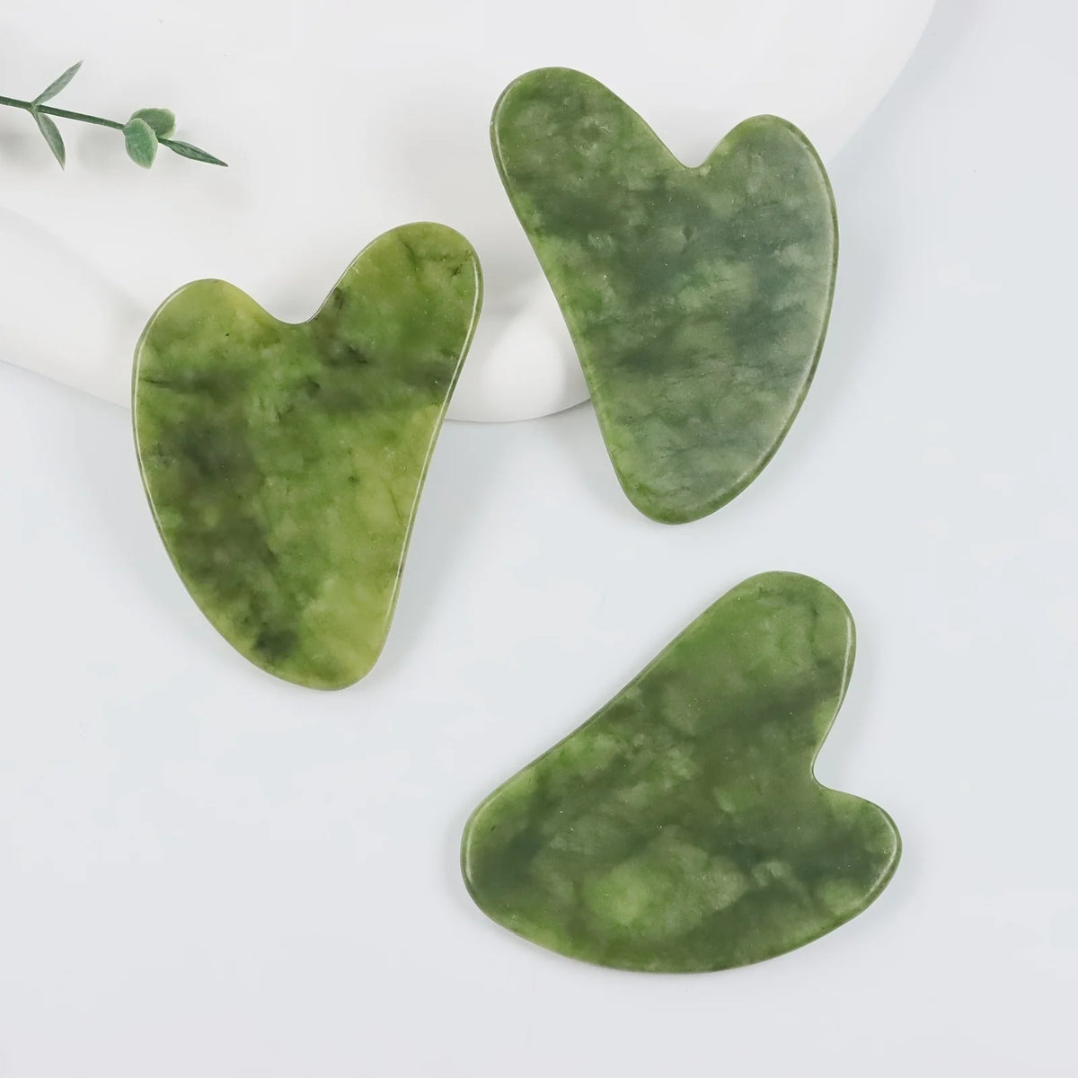 Three jade gua sha tools in various shades of green resting on a white surface.