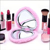 A heart-shaped foldable travel makeup mirror with a pink fuzzy background and makeup brushes.
