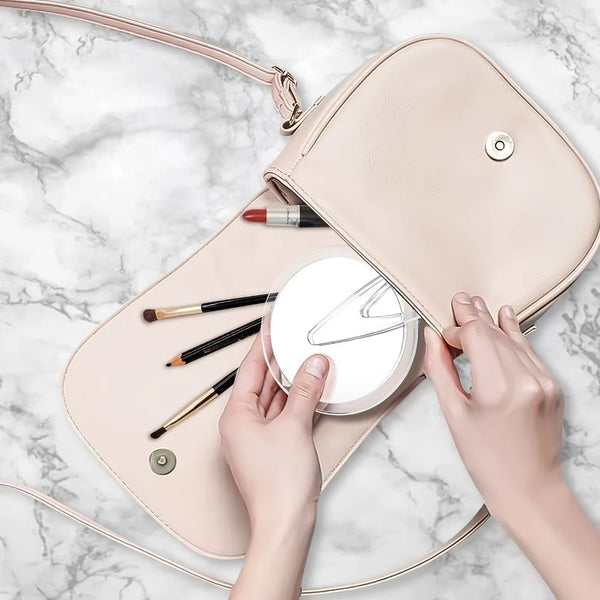 Hands hold a pink foldable vanity mirror with makeup brushes and a small round mirror on a marble surface.