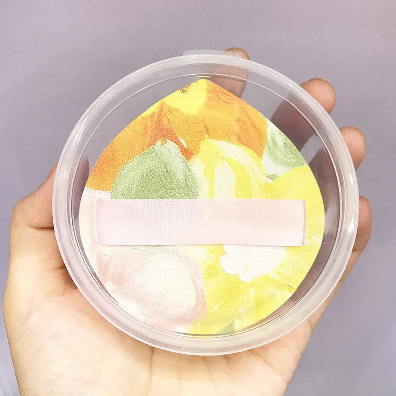 An air cushion makeup puff set with floral design is shown inside a clear storage box being held by a hand.
