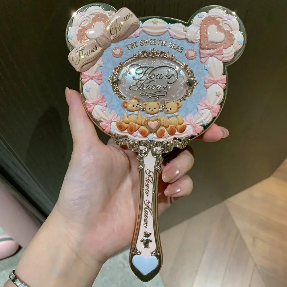 A hand holds a decorative makeup mirror set featuring pink and blue floral accents and small bear figures.