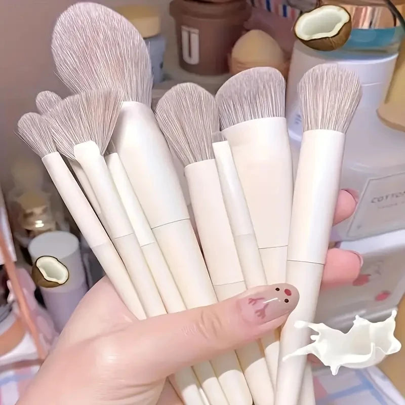 A hand holds a set of fourteen professional makeup brushes with light pink bristles and white handles.