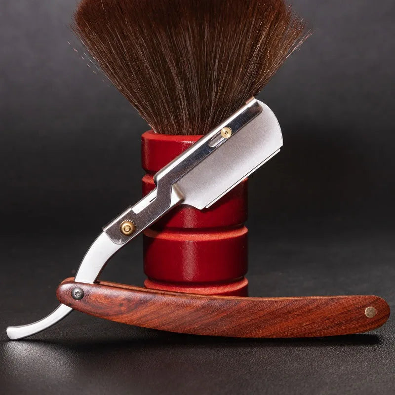 A professional shaving razor with a silver handle and wood grip rests on a red cylindrical stand, next to a shaving brush.