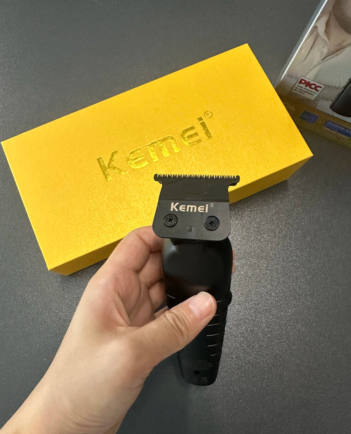 A hand holds a black Kemei electric hair clipper above a yellow box.