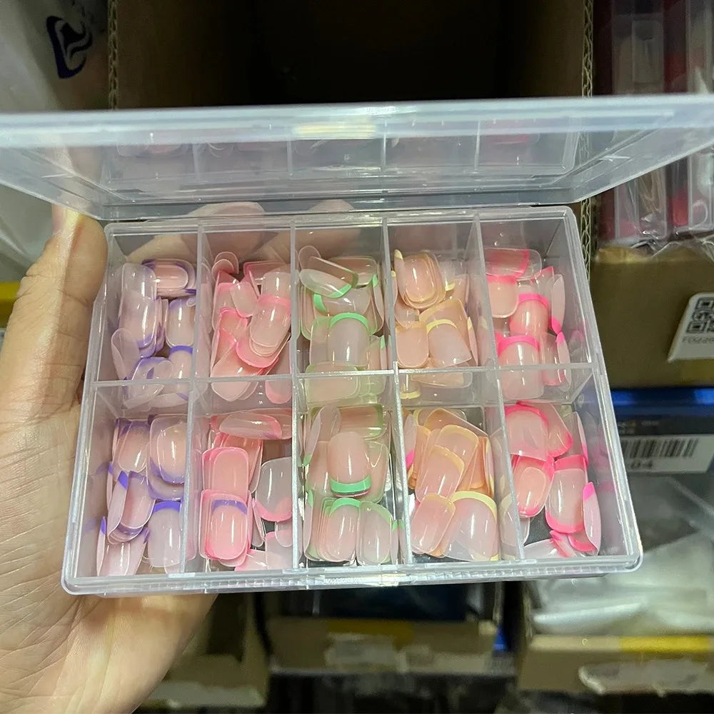 Une main tient une boîte en plastique transparent de faux ongles acryliques colorés dans différentes nuances de rose et de nu