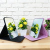 A foldable portable makeup mirror with a pink stand displaying yellow flowers against a white brick background.