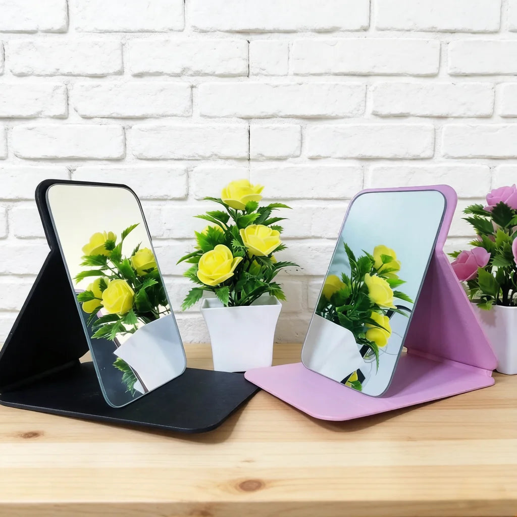 A foldable portable makeup mirror with a pink stand displaying yellow flowers against a white brick background.