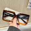 A hand holds a pair of brown-tinted square eyeglasses with clear lenses against a light background.