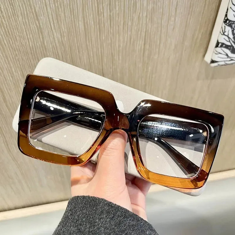 A hand holds a pair of brown-tinted square eyeglasses with clear lenses against a light background.