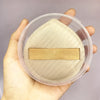 A hand holds a clear plastic storage box containing an air cushion makeup puff set with a beige pad and a tan strip.