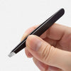 A hand holds a black precision stainless steel eyebrow tweezers with a silver tip.