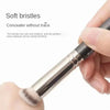 A hand is using a concealer brush to apply makeup, showing the soft bristles of the tool.