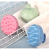 A pink silicone scalp massage brush and a blue silicone body brush are displayed on a white towel within a wire rack.
