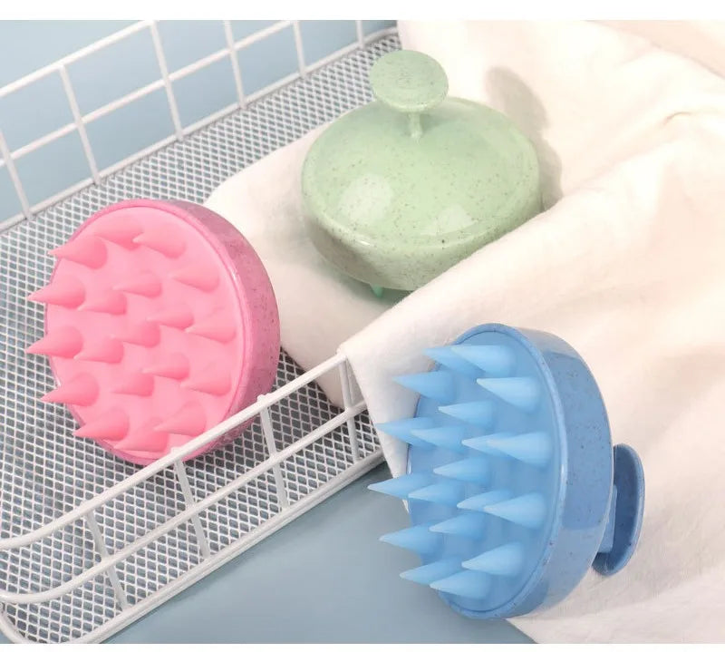 A pink silicone scalp massage brush and a blue silicone body brush are displayed on a white towel within a wire rack.