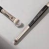 A double-ended eye shadow brush with a metal smudge tip and a fluffy blending shader on a gray background.