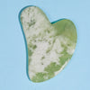 A smooth, heart-shaped piece of light green jade used as a Gua Sha tool for facial massage.