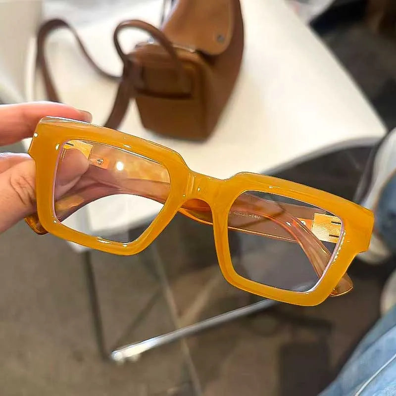 Une paire de lunettes de lecture carrées rétro teintées en jaune est tenue, avec un sac en cuir marron en arrière-plan.